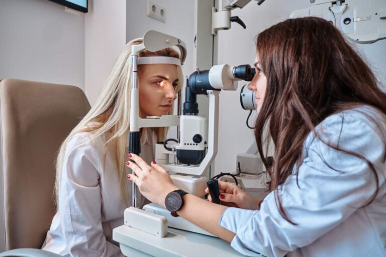 An optometrist performing a vision test with a patient reading a wall chart in a well-lit exam room.